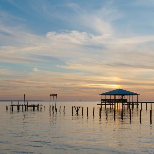 An image of a serene waterfront at sunset, featuring a dock with a small covered structure, and wooden posts reflecting in the calm water.