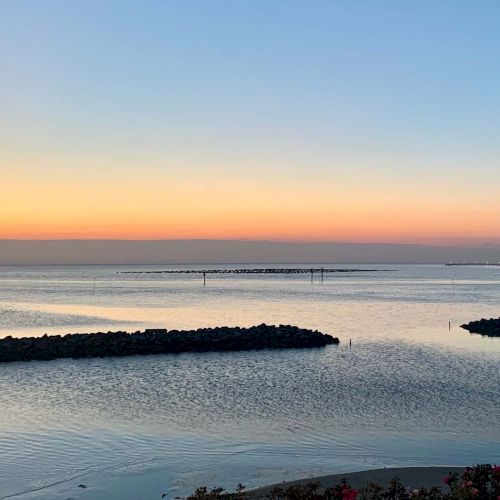 A serene sunset over a calm body of water, with rocky barriers and a distant tree line on the horizon. The sky is a gradient of warm colors.