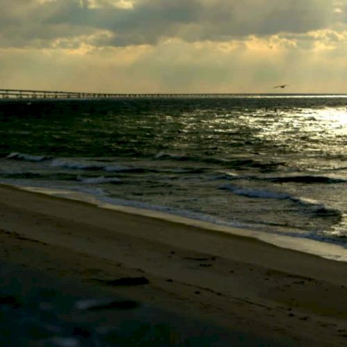 A beach scene during sunset with waves crashing on the shore and a long bridge extending into the horizon under a cloudy sky.