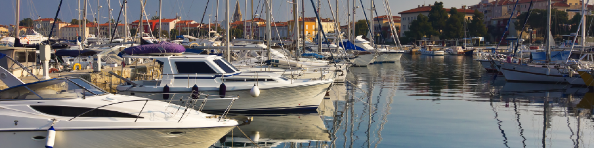 A marina with several boats docked, calm water reflecting the boats and clear sky, with buildings in the background ending the sentence.