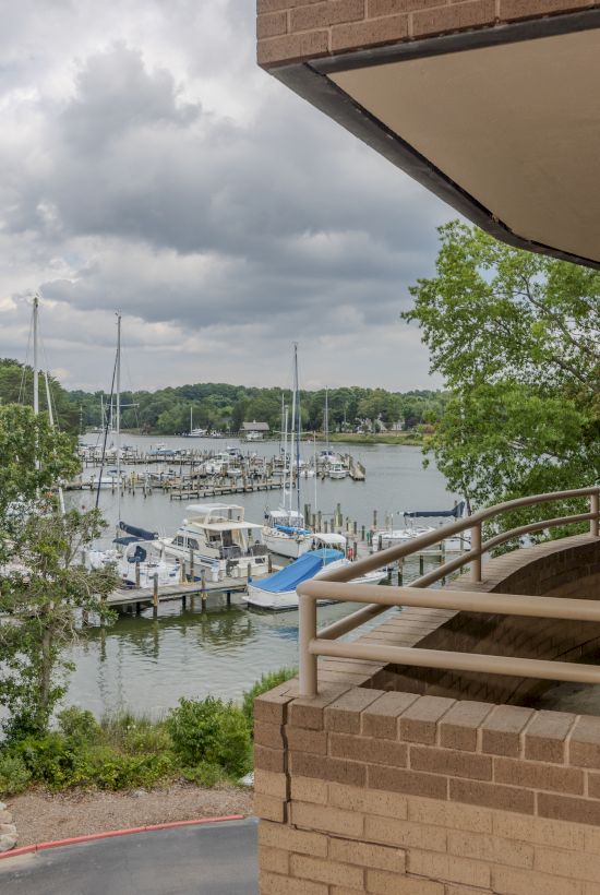 Image of a marina with sailboats, trees, and a cloudy sky, viewed from a balcony of a building with a brick facade and railing.