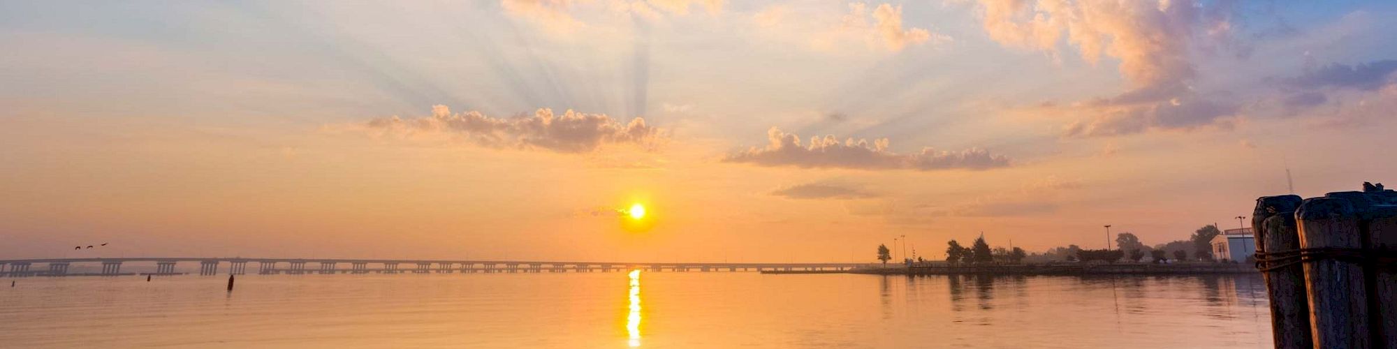 The image shows a serene sunset over calm waters with a bridge in the distance, and a peaceful shoreline with a few buildings and trees.