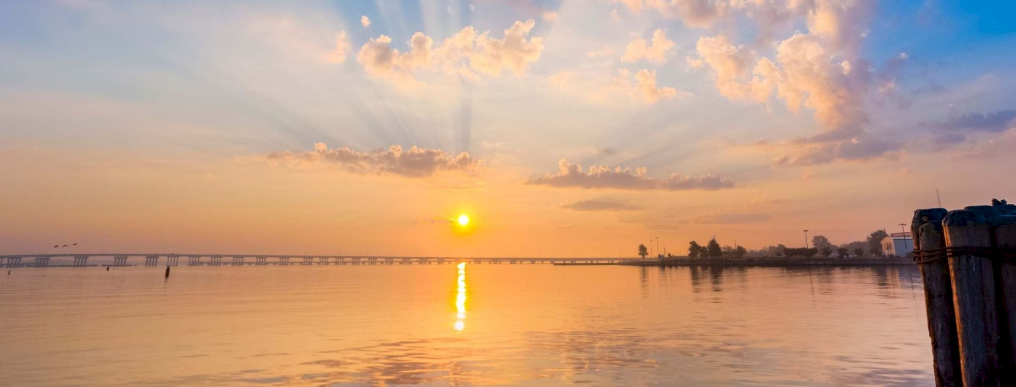 The image shows a serene sunset over calm waters with a bridge in the distance, and a peaceful shoreline with a few buildings and trees.